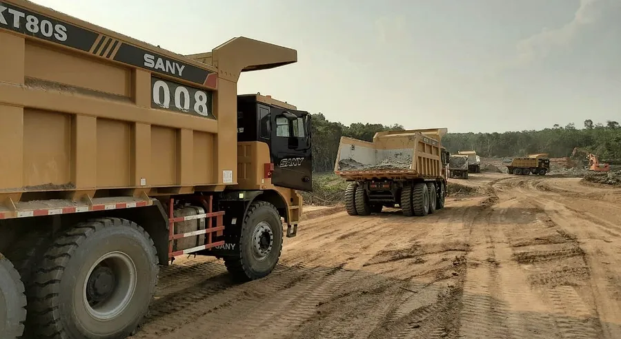 Loaded haul trucks moving in convoy along a graded laterite haul road cut through forest.