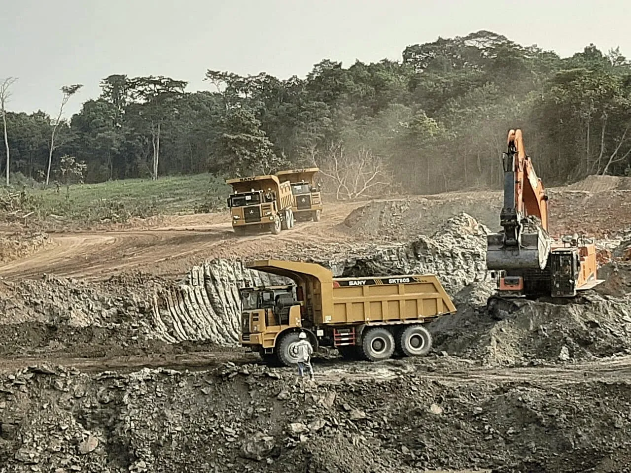 Excavator loading overburden into a haul truck at the edge of a pit, forest on the ridge behind.